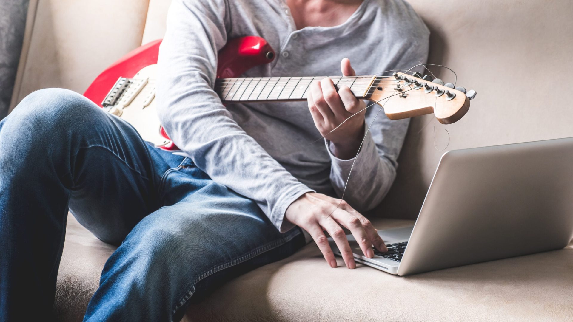 Casual young man playing guitar on sofa at home.